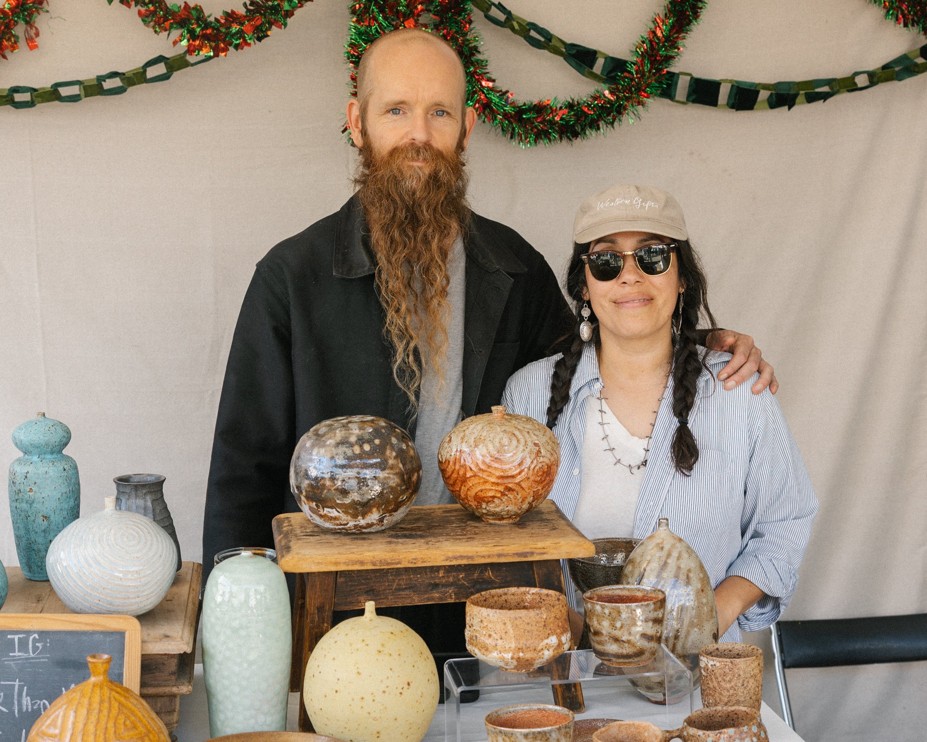 Two people standing behind a table with ceramic items at a market stall.