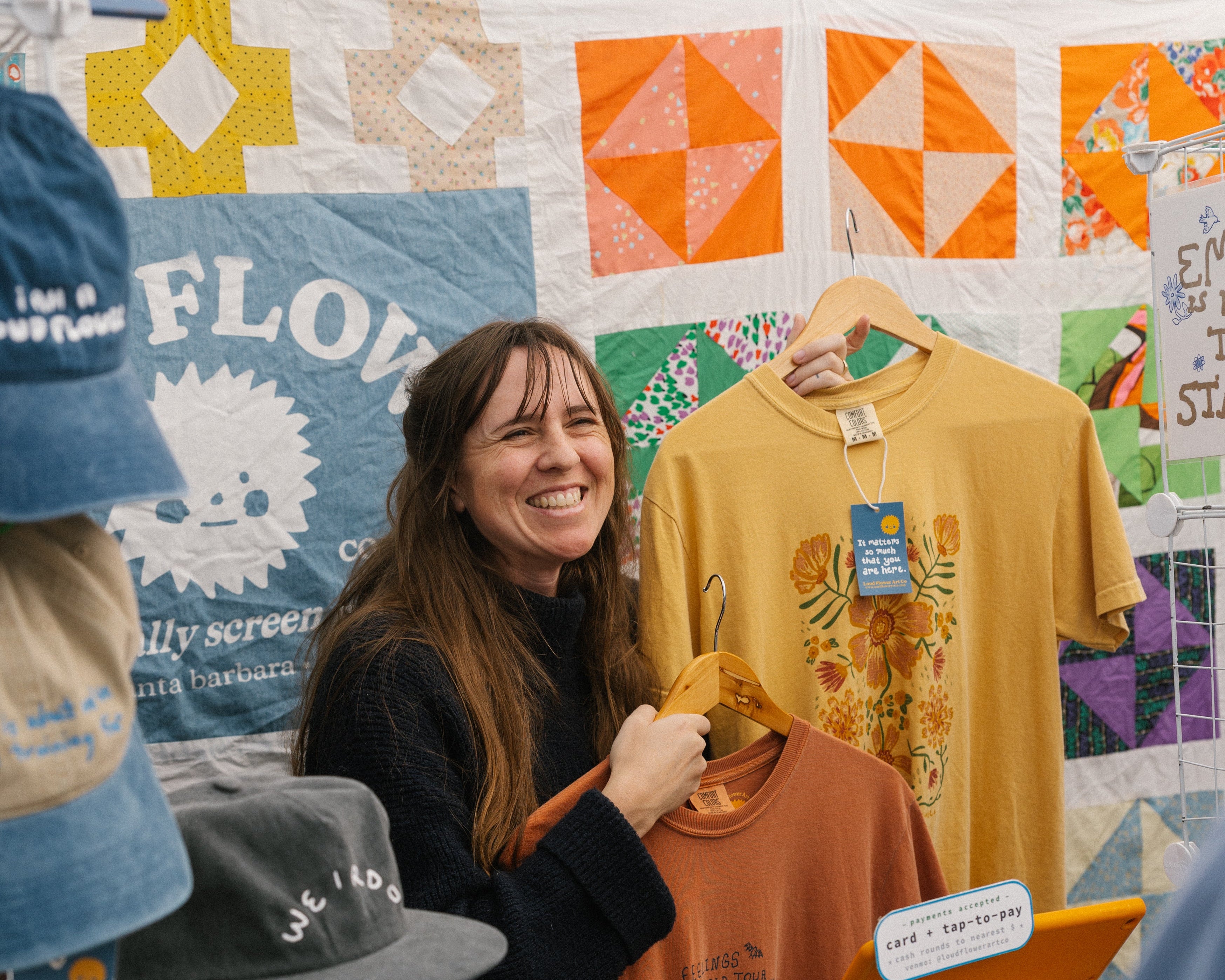 Woman holding a yellow t-shirt at a craft fair with various items on display.