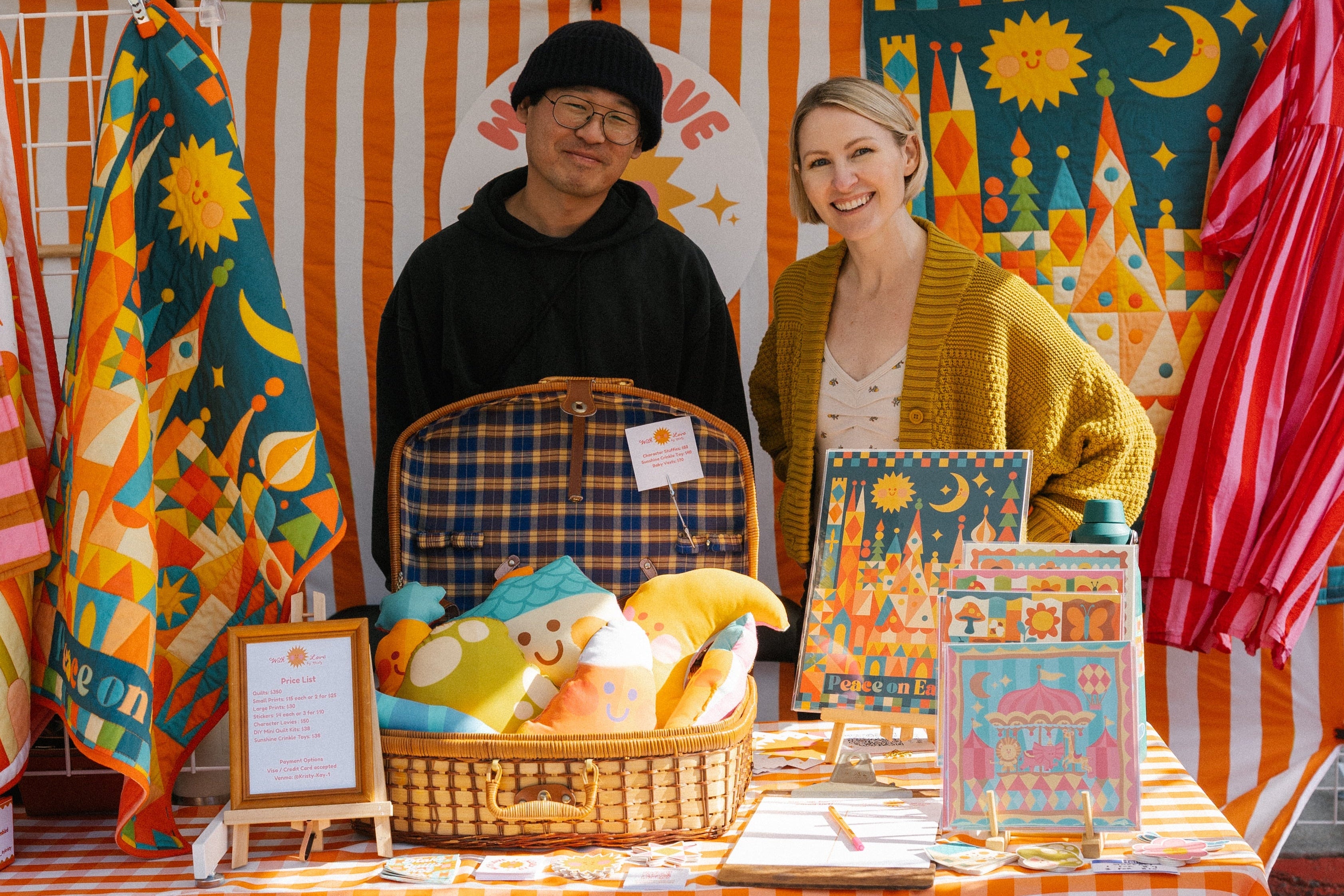 Two people standing behind a table with various items at an outdoor market stall.