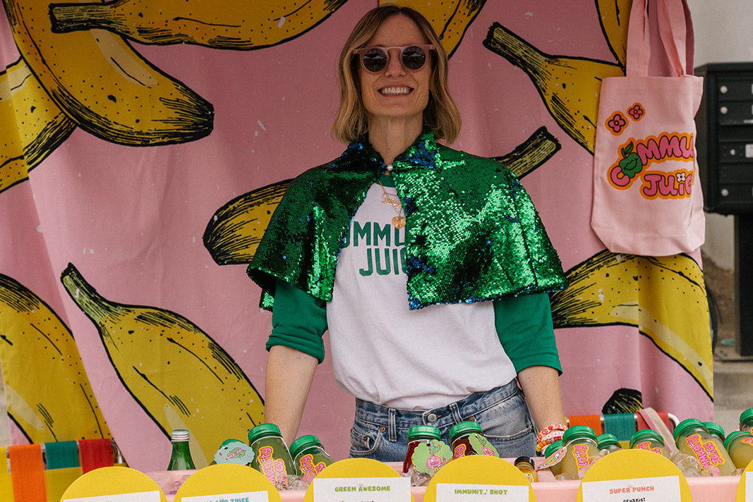 Person standing behind a colorful table with juice products, wearing a green sequin jacket and sunglasses, with a pink backdrop featuring bananas.
