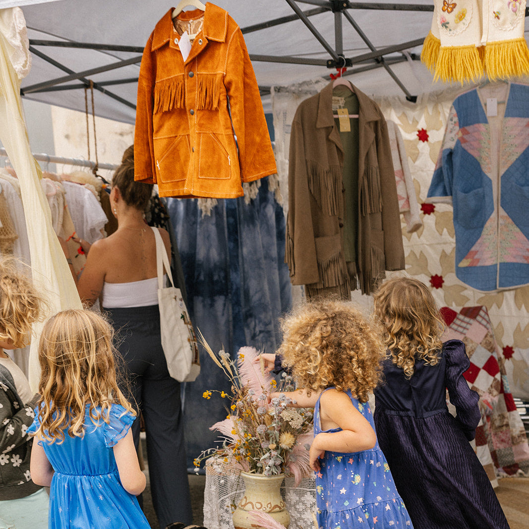 Children in blue dresses exploring a market stall with clothing and decor items.