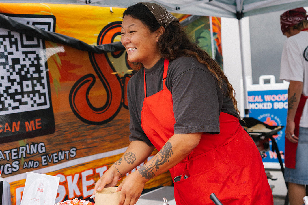 Woman in a red apron preparing food at an outdoor event under a striped canopy.
