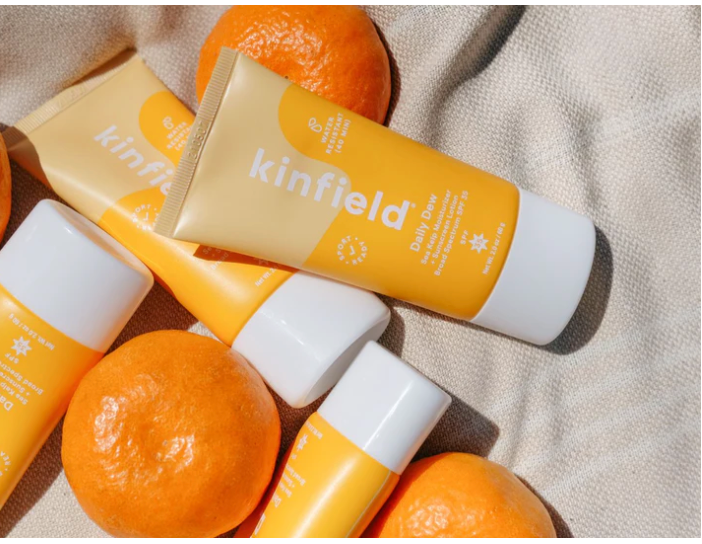 Top view of four orange and yellow Kinfield sunscreen bottles with oranges around them